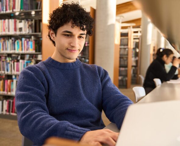 Man at a library looking at his laptop, with bookshelves in the background.