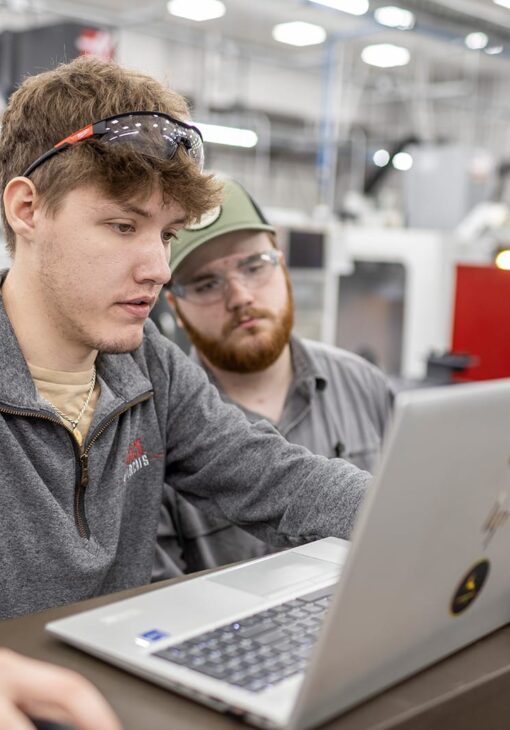 Two male students wearing safety glasses work together on a laptop in a technical lab, with industrial equipment visible in the background.
