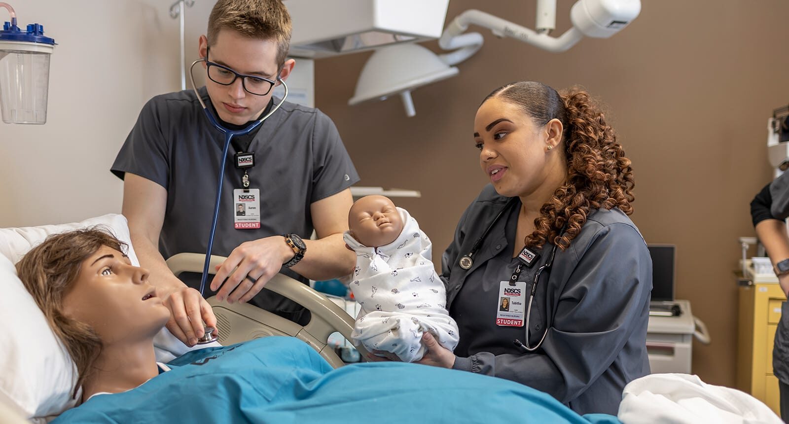 Three nursing students in scrubs practicing a childbirth scenario using a medical mannequin and a baby doll in a hospital simulation lab.
