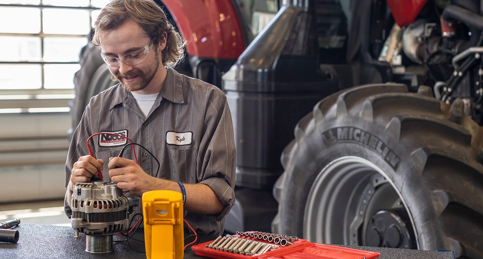 Male student using a multimeter to test an alternator on a workbench, with a red tractor and large Michelin tire in the background.