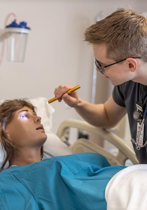 Nursing student using a penlight to examine the eye of a medical training mannequin during a clinical simulation.